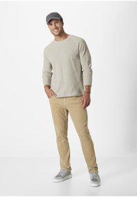 Beige Waffelstrick Langarmshirt, khakifarbene Cordhosen, graue Mütze und Sneakers mit grauen Akzenten. Das Modell steht vor einem weißen Hintergrund.