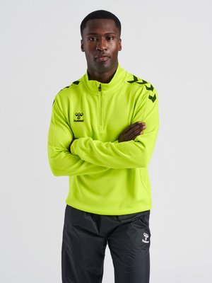 Mężczyzna ze skrzyżowanymi ramionami, ubrany w jaskrawożółtą bluzę na zamek z długim rękawem i czarne spodnie z logo marki na obu elementach odzieży.