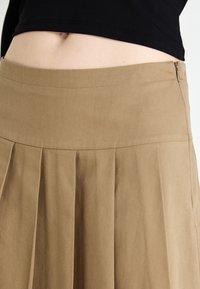 Une jupe plissée beige avec une taille lisse et une fermeture éclair sur le côté. Le tissu semble doux avec un léger éclat et une texture minimale.