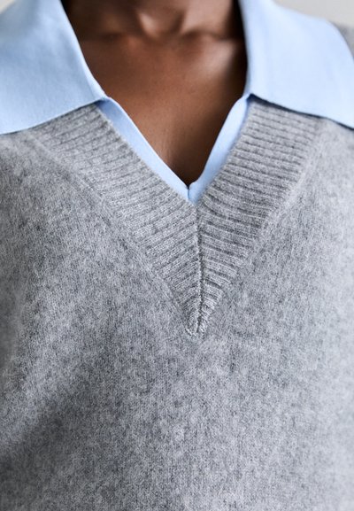 Jersey gris de punto con escote en V, hecho de material suave, con textura acanalada en el cuello. Camisa de collar azul claro debajo.