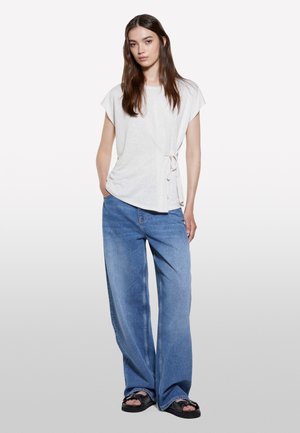 Haut blanc à manches courtes avec détail de nœud sur le côté, associé à un jean bleu clair à jambe large et des sandales noires. Tissu lisse et coupe décontractée.