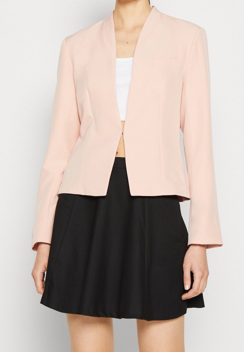 Femme portant un blazer rose clair sur un crop top blanc et une jupe noire taille haute, montrée des épaules jusqu'à mi-cuisse.
