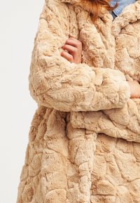 Tan, konstläderjacka med en texturerad, plyschyta som har ett skrynkligt mönster. Designen inkluderar en sjalkrage och synliga sömmar.