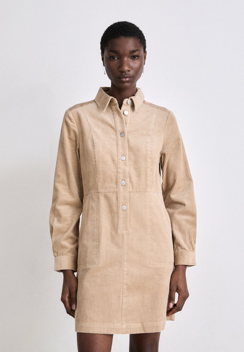 Beige Kordkleid mit Kragen, Knopfleiste, langen Ärmeln und zwei vorderen Taschen mit gerippter Textur.