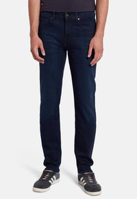 Jeans in denim blu scuro con vestibilità slim, tasche frontali e chiusura a bottoni classica. Abbinati a sneaker grigie con strisce bianche.