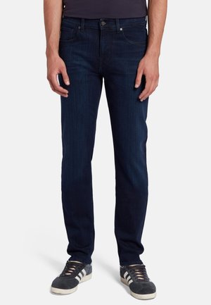 Jeans en denim bleu foncé avec une coupe slim, poches avant et fermeture à bouton classique. Associés à des baskets grises avec des rayures blanches.