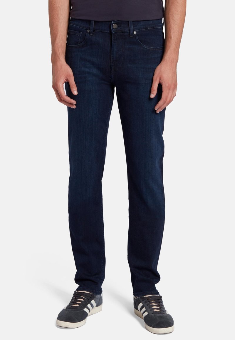 Jeans in denim blu scuro con vestibilità slim, tasche frontali e chiusura a bottoni classica. Abbinati a sneaker grigie con strisce bianche.