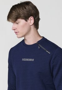 Joven con cabello castaño que lleva una sudadera acolchada color azul marino con el texto "KOROSHI" y un detalle de cremallera cerca del cuello.