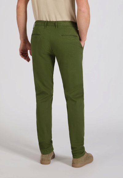 Man staat met handen in de zakken, draagt olijfgroene slim-fit broek en beige suède schoenen tegen een effen achtergrond.