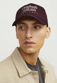 Gorra de béisbol burdeos con "Originals Atelier EST. 1990" bordado en blanco. La tela es suave, con visera curvada y banda ajustable en la parte trasera.