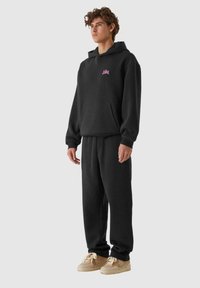 Set di felpa nera con pantaloni tuta realizzato in materiale morbido, con tasca frontale, polsini a coste e un piccolo logo rosa sul petto.