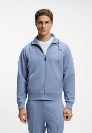 Uomo che indossa una felpa con cappuccio azzurro chiaro con cerniera e pantaloni coordinati, con una maglietta bianca sotto, in piedi davanti a uno sfondo bianco.