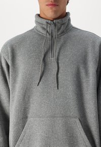 Sudadera de forro polar gris con un alto cuello, cierre de media cremallera al frente, cordones y un bolsillo canguro en la parte delantera. Textura suave, patrón jaspeado sutil.