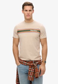 Camiseta de algodón color beige con un diseño de rayas multicolores en verde, naranja y azul, que presenta el logo de "SuperDry". Se lleva con una camisa de cuadro atada a la cintura.