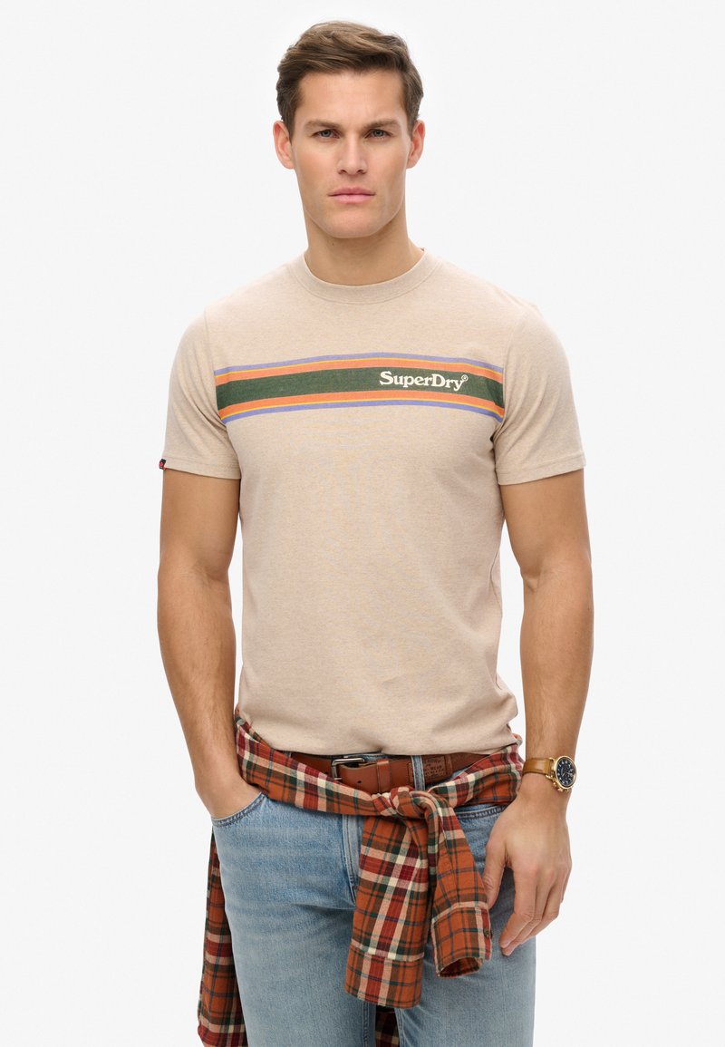 Camiseta de algodón color beige con un diseño de rayas multicolores en verde, naranja y azul, que presenta el logo de "SuperDry". Se lleva con una camisa de cuadro atada a la cintura.