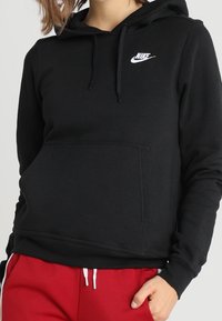 Svart Nike-hoodie med framficka, huva med dragsko och vit logga. Har långa ärmar och ett mjukt, texturerat tyg.