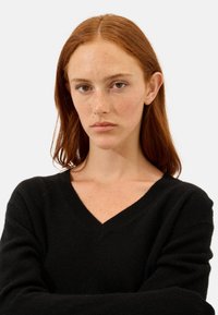Schwarzer V-Ausschnitt-Pullover mit langen Ärmeln, aus weichem Stoff gefertigt. Strukturierte Optik mit gerippten Abschlüssen, getragen von einem Modell mit rotem Haar.