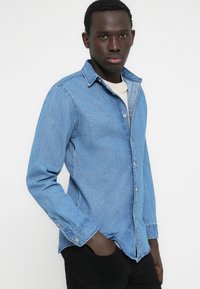 Hellblaues Jeanshemd mit durchgehender Knopfleiste, Kragen, langen Ärmeln und leicht verwaschener Textur. Kombiniert mit schwarzen Hosen.