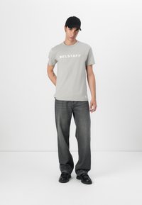 Γκρι βαμβακερό t-shirt με το "BELSTAFF" σε λευκό χρώμα, συνδυασμένο με χαλαρές σκούρες γκρι τζιν και μαύρα παπούτσια. Το μοντέλο φοράει ένα μαύρο καπέλο.