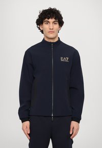 Mornarska zip-up jakna z visokim ovratnikom, črnimi stranskimi paneli, zlatim vezenim logotipom na prsih, prilegajočimi manšetami in mehkimi tkaninami.