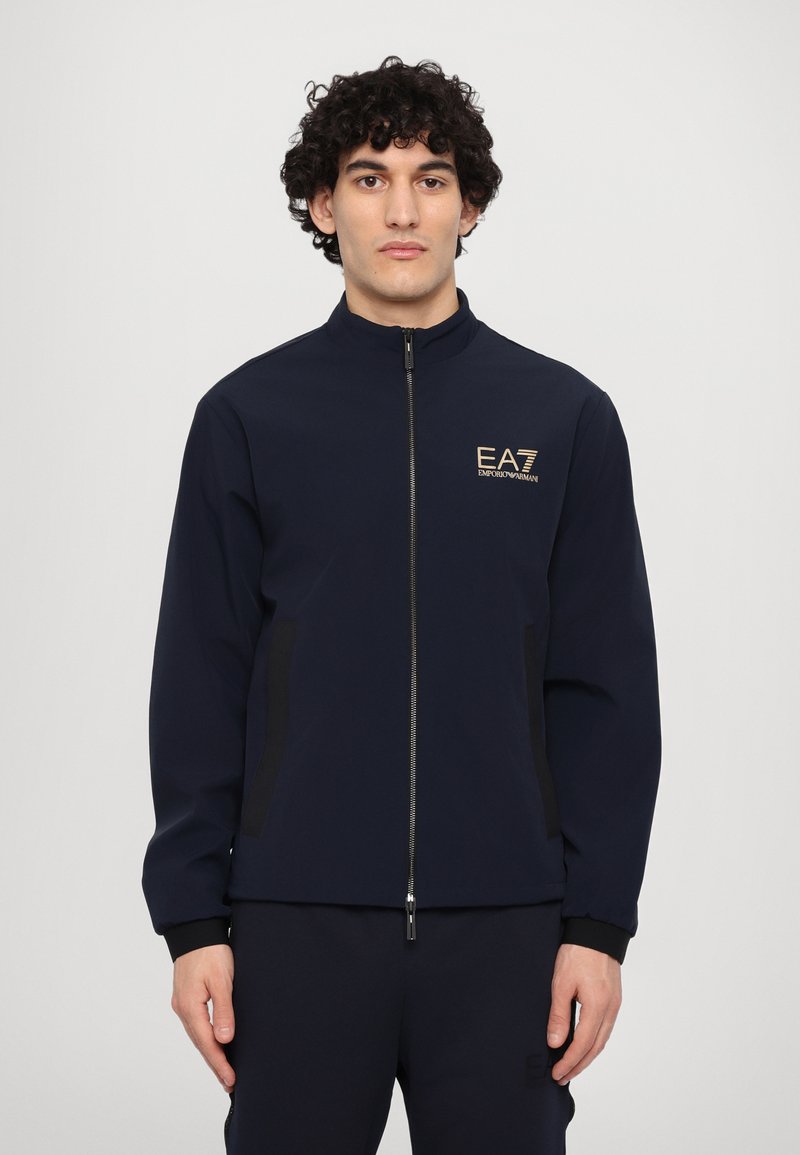 Mornarska zip-up jakna z visokim ovratnikom, črnimi stranskimi paneli, zlatim vezenim logotipom na prsih, prilegajočimi manšetami in mehkimi tkaninami.