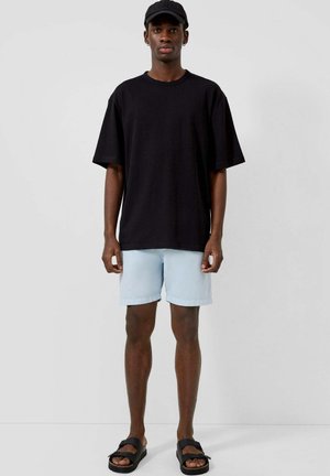 Młody mężczyzna stojący, ubrany w czarną czapkę, oversize czarny T-shirt, jasnoniebieskie szorty i czarne sandały na jednolitym tle.