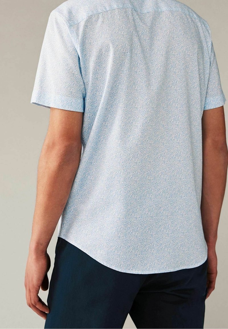 Homme portant une chemise bleu clair à manches courtes avec un motif floral subtil et un pantalon foncé, debout dos à la caméra.