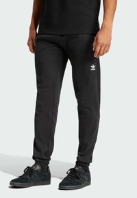 Sorte ribbede Adidas joggingbukser med hvidt logo, tætsiddende design, elastiske manchetter, parret med sorte sneakers med tre striber og snørebånd.