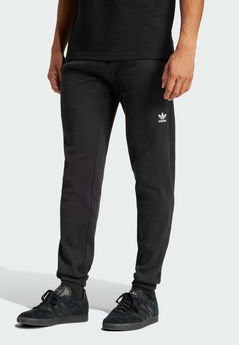 Sorte ribbede Adidas joggingbukser med hvidt logo, tætsiddende design, elastiske manchetter, parret med sorte sneakers med tre striber og snørebånd.
