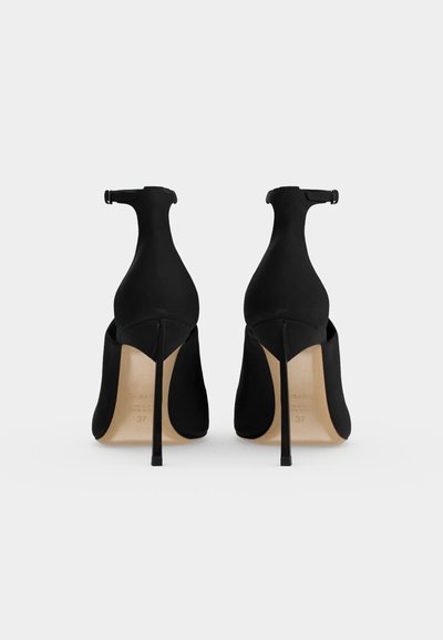 Paire de chaussures à talons hauts noires avec brides à la cheville, vues de dos sur un fond uni clair.