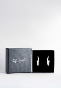 Silverörhängen med eleganta kurvor, som ligger mot en svart sammetsbakgrund i en texturerad svart låda. Kännetecknas av klara ädelstenar inlagda i designen.