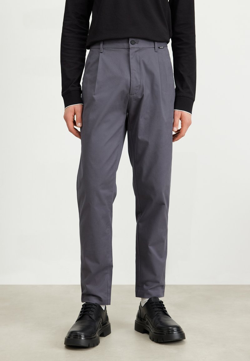 Grey tailored pants with a smooth texture, featuring a front crease, button closure, and side pockets, paired with black shoes.