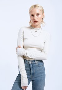 Light beige long-sleeve top with thumbholes, fitted design, and soft texture, paired with high-waisted blue jeans. Multiple delicate necklaces.