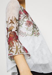 Blusa semitranslúcida blanca con bordado floral en rojo, verde y dorado. Tiene mangas acampanadas y un diseño fluido.