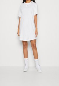 Bílá bavlněná šatová tričko s krátkým rukávem, s černým logem Nike na hrudi. Tato kombinace je doplněna bílými ponožkami Nike a teniskami.