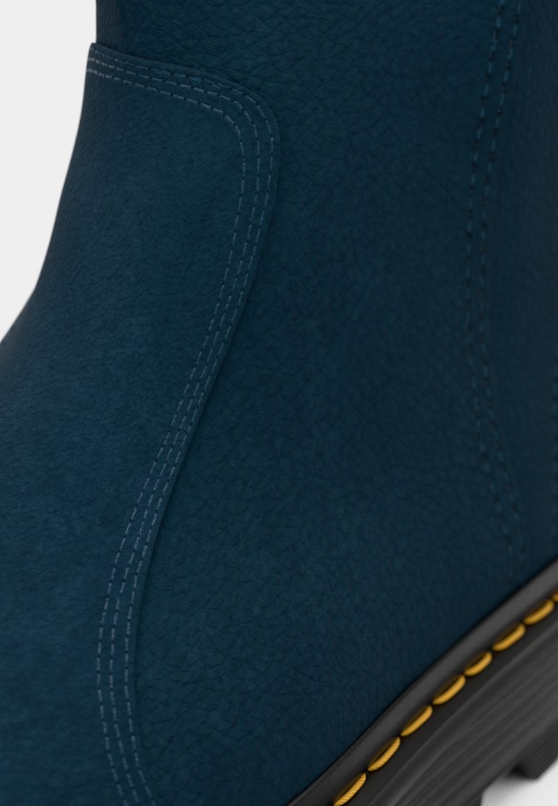 Botte bleue texturée avec des détails de couture visibles, dotée d'une semelle noire et de surpiqûres jaunes contrastantes le long du bord de la semelle.