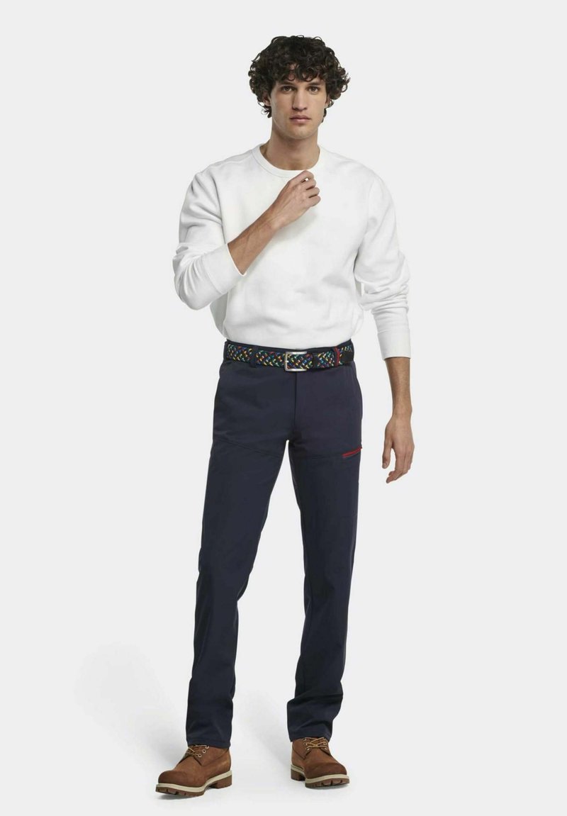 Weißer Kapuzenpullover, navyfarbene Hose mit einem gemusterten Gürtel, braune Stiefel. Die Hose hat rote Akzente und Taschen. Das Modell steht mit den Händen nah am Brustkorb.