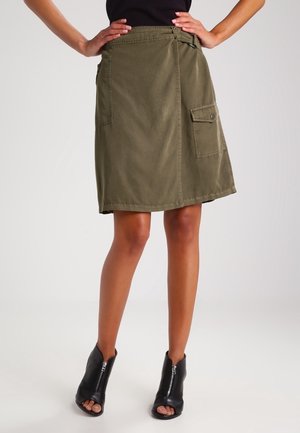 Femme portant une jupe longue jusqu'aux genoux couleur vert olive avec rabat de poche avant et des bottines noires à bouts ouverts avec fermetures éclair devant.