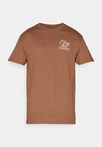 Hnědý bavlněný tričko s krátkým rukávem, kulatým výstřihem a bílým logem "Obey Studios" na levém hrudníku. Hladká textura, neformální design.