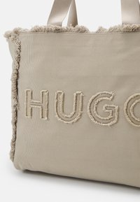 Beige Leinencanvas-Tasche mit ausgefransten Kanten und eingeprägtem "HUGO"-Schriftzug in einem strukturierten Faden. Verfügt über stabile, breite Stoffträger.