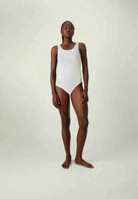 Γυναίκα που στέκεται ξυπόλητη, φορώντας μια αμάνικη λευκή φόρμα, μπροστά σε μια απλή ανοιχτόχρωμη φόντο, κοιτάζοντας μπροστά με χαλαρωμένα χέρια.