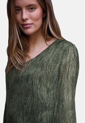 Joven mujer con cabello largo castaño claro que lleva una blusa verde con estampado y mangas abullonadas de tela transparente, posando frente a un fondo liso.