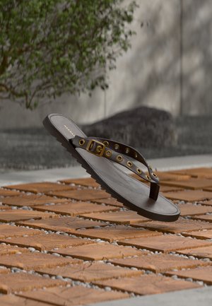 Sandale en cuir marron avec œillets dorés et boucle flottant au-dessus de carreaux de pierre orange et de galets avec un arbre et un mur gris en arrière-plan.