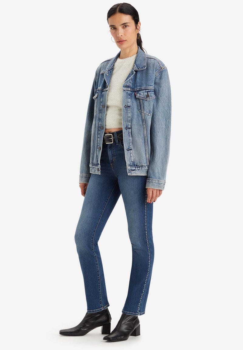 Veste en denim bleu clair avec une coupe décontractée, associée à un pull crème et un jean bleu slim. Des bottines noires à talons carrés complètent le look.