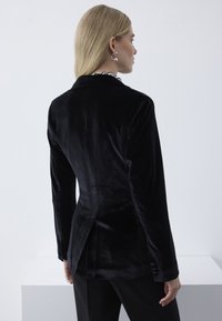 Kvinde med glat blondt hår iført sort fløjlsblazer samt sølvfarvet kuglehalskæde og øreringe, set bagfra mod en ensartet hvid baggrund.