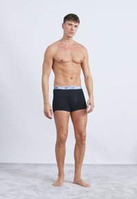 Černé boxerky Nike s bílým pasem a logem. Model stojí naboso na šedém koberci a ukazuje slim-fit střih.