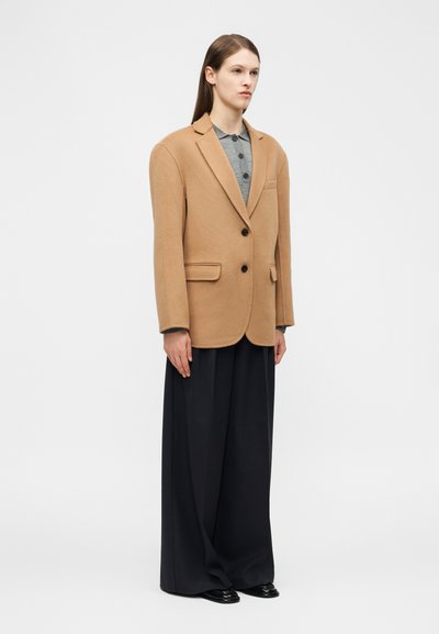 Egyenes hajú nő túlméretezett tevebarna blézerben, szürke galléros ingben, széles fekete nadrágban és fekete cipőben, fehér háttér előtt áll.