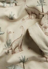 Bomullstyg med ett mönster av ljusbruna dinosaurier och grön vegetation på en krämfärgad bakgrund, med en mjuk och strukturerad yta.