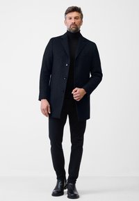 Dunkler Wollmantel mit Reverskragen, drei Knöpfen und einer Brusttasche, kombiniert mit schwarzen Slim-Fit-Hosen und glänzenden schwarzen Ankle Boots.