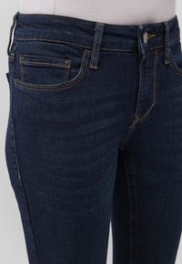 Dark blue denim jeans with a slim fit, featuring five pockets, orange stitching, and a high waistband. Smooth texture and classic design.